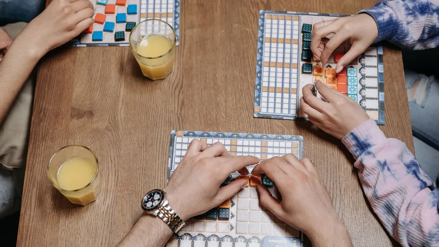Hands around a wooden table placing colourful tiles — the kind of table game ChampionChoice builds for the browser